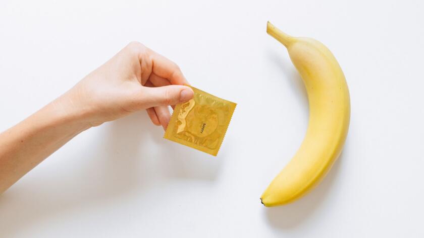 A hand holding a condom next to a banana on a white background, symbolizing safe sex education.