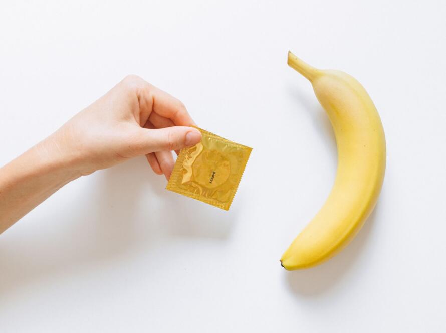 A hand holding a condom next to a banana on a white background, symbolizing safe sex education.