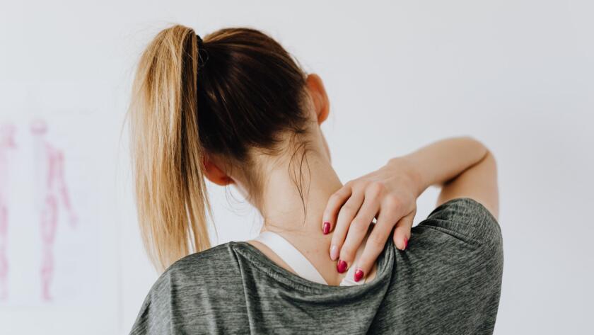 Back view of slim blond female in casual wear with manicure indicating pain spot on back while sitting in front of white