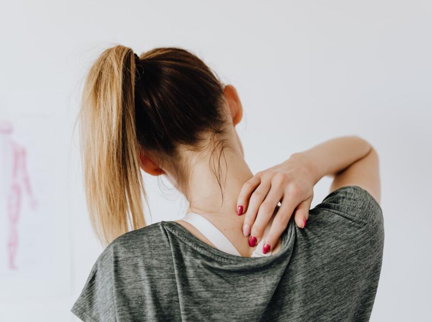 Back view of slim blond female in casual wear with manicure indicating pain spot on back while sitting in front of white