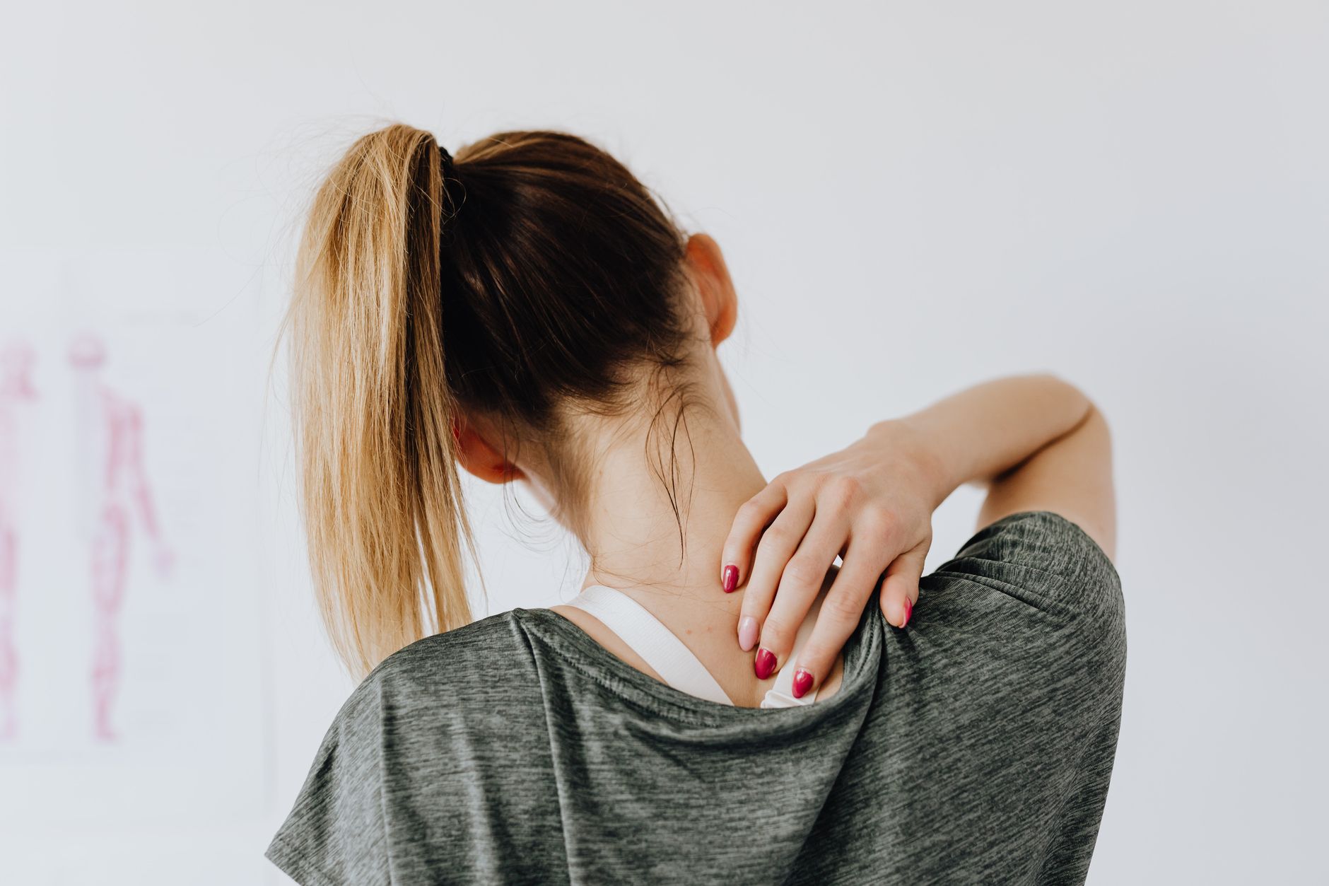 Back view of slim blond female in casual wear with manicure indicating pain spot on back while sitting in front of white